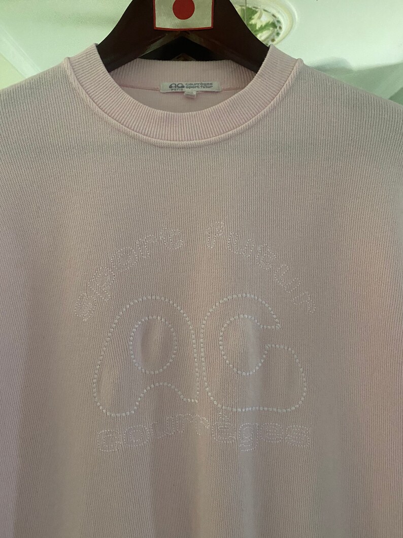 Puede incluir: Una camisa de manga larga de color rosa claro con un logotipo bordado en blanco que dice "Comme des Gar&ccedil;ons Play" y presenta un coraz&oacute;n con un contorno cosido en blanco.