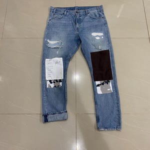 Puede incluir: Vaqueros de mezclilla azul claro con detalles desgastados, incluyendo rasgaduras y parches. Los vaqueros presentan un parche de gamuza marrón y otros parches de tela con diseños abstractos. Los puños están doblados.