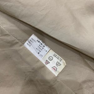 K&ouml;nnte beinhalten: Nahaufnahme eines beigefarbenen Stoffes mit einem angebrachten Etikett. Das Etikett enth&auml;lt japanischen Text und Symbole, einschlie&szlig;lich Waschanweisungen. Der Stoff scheint eine Mischung aus Materialien zu sein, darunter Wolle, Kaschmir und Nylon.