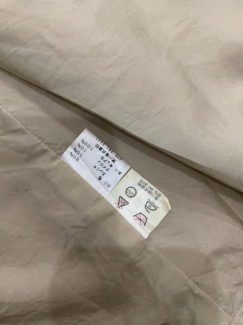 K&ouml;nnte beinhalten: Nahaufnahme eines beigefarbenen Stoffes mit einem angebrachten Etikett. Das Etikett enth&auml;lt japanischen Text und Symbole, einschlie&szlig;lich Waschanweisungen. Der Stoff scheint eine Mischung aus Materialien zu sein, darunter Wolle, Kaschmir und Nylon.