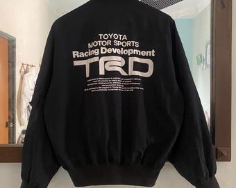 May include: Black bomber jacket with a white Toyota Motor Sports Racing Development TRD logo on the back. The jacket has a zipper closure and ribbed cuffs and hem.