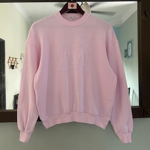 Puede incluir: Una sudadera rosa con un logotipo bordado en blanco que dice "A Cold Wall*" y tiene un dise&ntilde;o de nube.