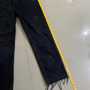 Puede incluir: Vaqueros negros de denim desgastado con dobladillos deshilachados. Los vaqueros se miden con una cinta m&eacute;trica amarilla.