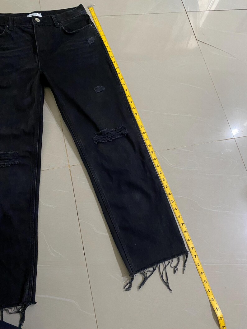 Puede incluir: Vaqueros negros de denim desgastado con dobladillos deshilachados. Los vaqueros se miden con una cinta m&eacute;trica amarilla.