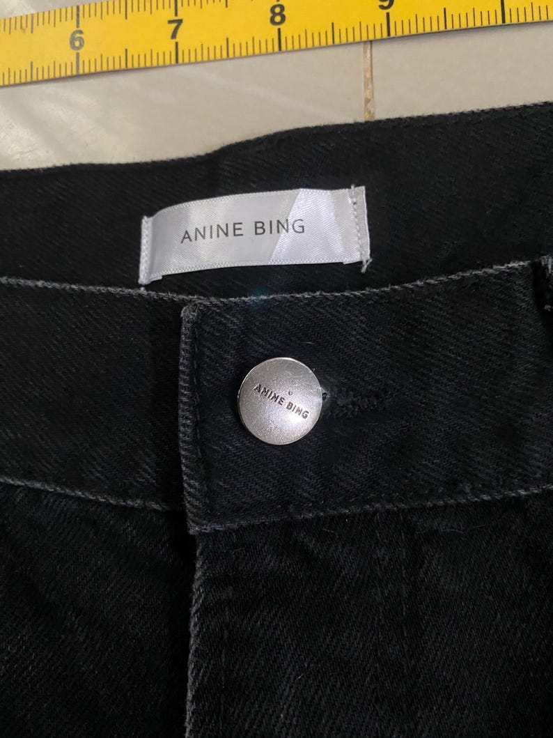 Puede incluir: Vaqueros negros de denim con una etiqueta blanca que dice "Anine Bing" y un bot&oacute;n plateado con el mismo texto.