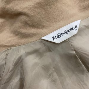 K&ouml;nnte beinhalten: Nahaufnahme eines beigefarbenen Kleidungsst&uuml;cks mit Satinfutter. Ein wei&szlig;es Etikett mit dem Text "Yves Saint Laurent" ist sichtbar. Das Kleidungsst&uuml;ck scheint ein Mantel oder eine Jacke mit weicher, strukturierter Oberfl&auml;che zu sein.