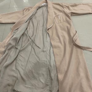 K&ouml;nnte beinhalten: Ein beiger Bademantel mit wei&szlig;er Paspelierung und passendem G&uuml;rtel. Der Bademantel hat eine Tasche mit einem Logo auf der Vorderseite. Das Innenfutter ist hellbeige. Der Bademantel liegt flach auf einer hellen Oberfl&auml;che.