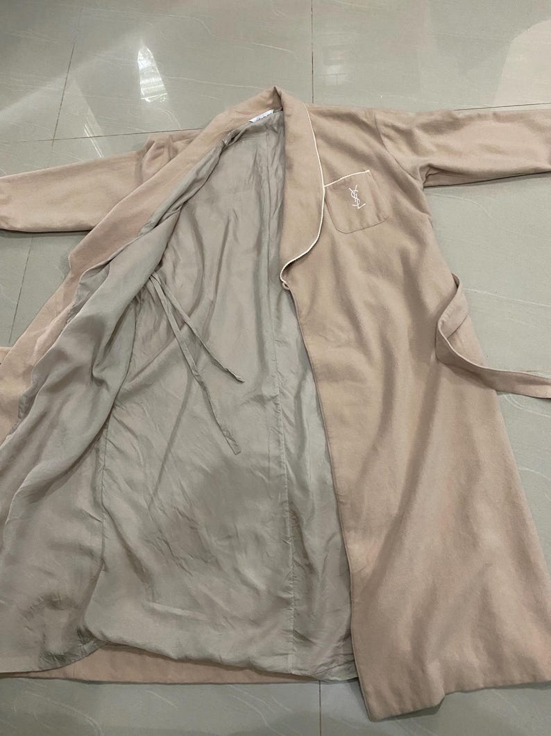 K&ouml;nnte beinhalten: Ein beiger Bademantel mit wei&szlig;er Paspelierung und passendem G&uuml;rtel. Der Bademantel hat eine Tasche mit einem Logo auf der Vorderseite. Das Innenfutter ist hellbeige. Der Bademantel liegt flach auf einer hellen Oberfl&auml;che.