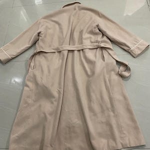 K&ouml;nnte beinhalten: Ein beigefarbener Trenchcoat mit passendem G&uuml;rtel, flach auf einer gefliesten Oberfl&auml;che liegend. Der Mantel hat lange &Auml;rmel mit wei&szlig;em Besatz an den Manschetten und einen wei&szlig;en Kragen. Der Mantel ist neutral gef&auml;rbt.