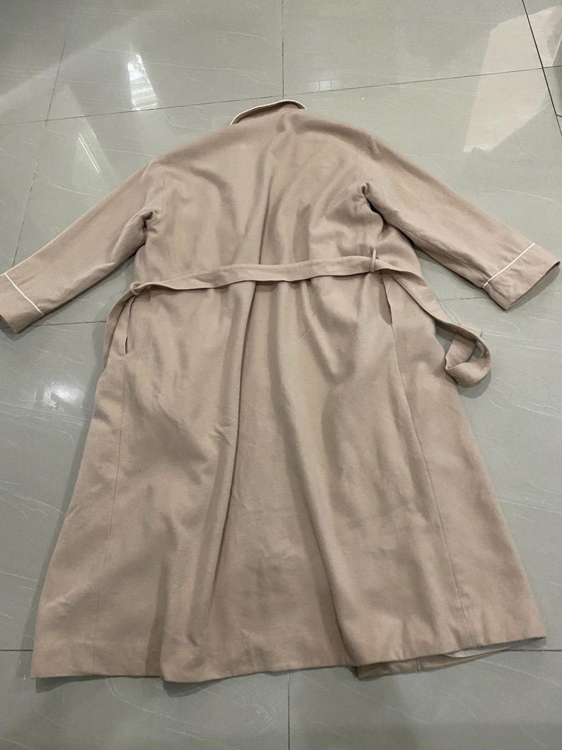 K&ouml;nnte beinhalten: Ein beigefarbener Trenchcoat mit passendem G&uuml;rtel, flach auf einer gefliesten Oberfl&auml;che liegend. Der Mantel hat lange &Auml;rmel mit wei&szlig;em Besatz an den Manschetten und einen wei&szlig;en Kragen. Der Mantel ist neutral gef&auml;rbt.