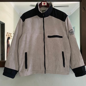Puede incluir: Una chaqueta de forro polar gris con detalles negros y cierre de cremallera. La chaqueta tiene un parche negro en la manga derecha con el texto "Cabela's Outdoor World" y "Polartec" en blanco.