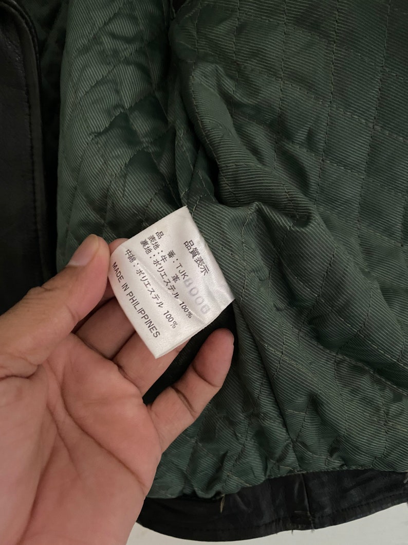 May include: A white tag with black text that reads "品質表示 番:TJKB006 表地:牛革 裏地:ポリエステル 100% 中綿:ポリエステル 100% MADE IN PHILIPPINES".