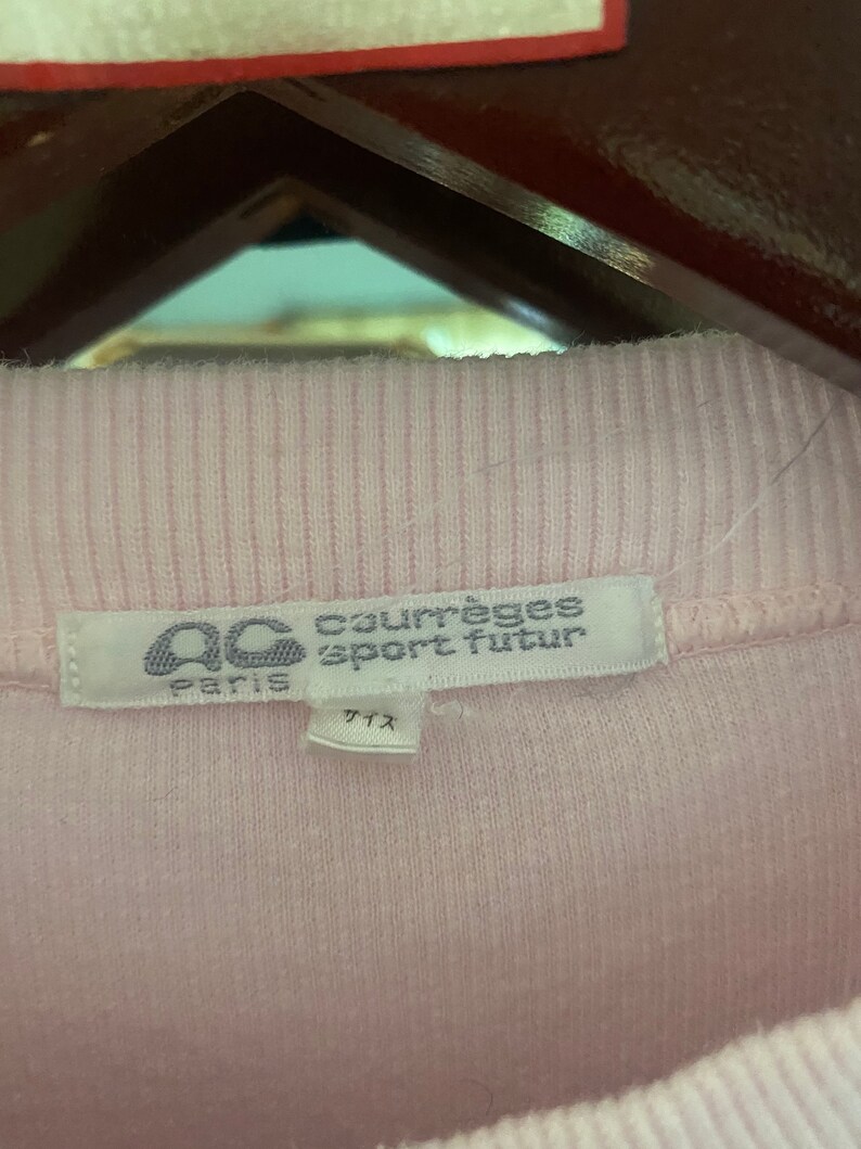 Puede incluir: Una camiseta de punto de canal&eacute; rosa con una etiqueta blanca que dice "Courr&egrave;ges Sport Futur Paris" y "サイズ" en japon&eacute;s.