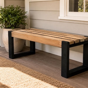 Può includere: Una panca rettangolare con seduta composta da listelli in legno marrone chiaro e una struttura robusta formata da spesse gambe in metallo nero opaco. Il design presenta una superficie superiore piatta e a doghe sostenuta da due basi in metallo nero.