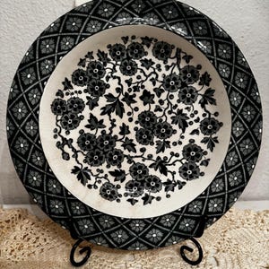 May include: A round ceramic plate featuring a black and white floral pattern in the center and a black and white diamond lattice border with small flower accents. The plate has a glossy finish and is displayed on a black metal stand.