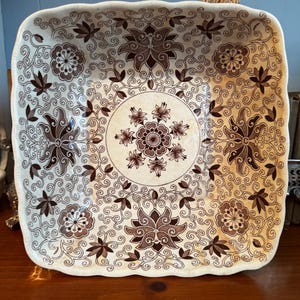 May include: A square ceramic bowl with scalloped edges, featuring a cream-colored base decorated with a dark brown intricate floral pattern. The design includes a central circular medallion surrounded by scrolling vines, leaves, and blossoms.
