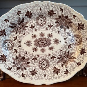 May include: An oval ceramic serving platter with a scalloped edge, featuring a cream-colored base decorated with a dark brown transferware pattern of intricate floral motifs, stylized blossoms, and delicate scrolling vine filigree.