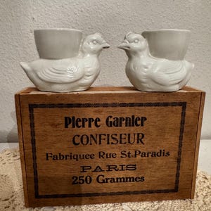May include: Two white ceramic bird-shaped egg cups resting on a rectangular wooden box. The box features black printed text reading: Pierre Garnier CONFISEUR Fabriquee Rue St. Paradis PARIS 250 Grammes, enclosed within a decorative black border.