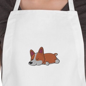 May include: White kitchen apron with a cartoon image of a brown and white corgi dog sleeping on it.