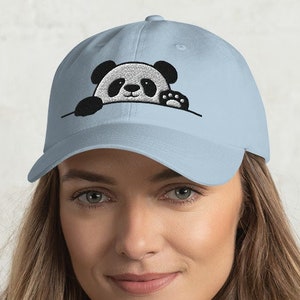 Può includere: Un berretto da baseball blu chiaro con un panda ricamato che sporge dal bordo. Il panda è nero e bianco con un'espressione carina.