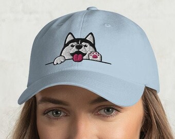Könnte beinhalten: Hellblaue Baseballkappe mit einem gestickten Husky-Hund-Design. Der Hund hat schwarz-weißes Fell, eine rosa Zunge und eine Pfote, die über den Rand schaut. Ein stilvolles und bequemes Accessoire für Hundeliebhaber.