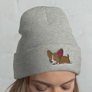 May include: A gray beanie with a cute cartoon dog embroidered on the front. The dog is brown with pink ears and is lying down.