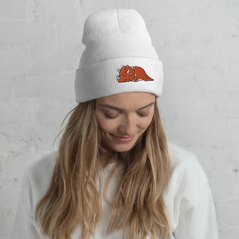 Puede incluir: Gorro de punto blanco con un dise&ntilde;o de triceratops bordado en marr&oacute;n. El triceratops est&aacute; acostado con los ojos cerrados.