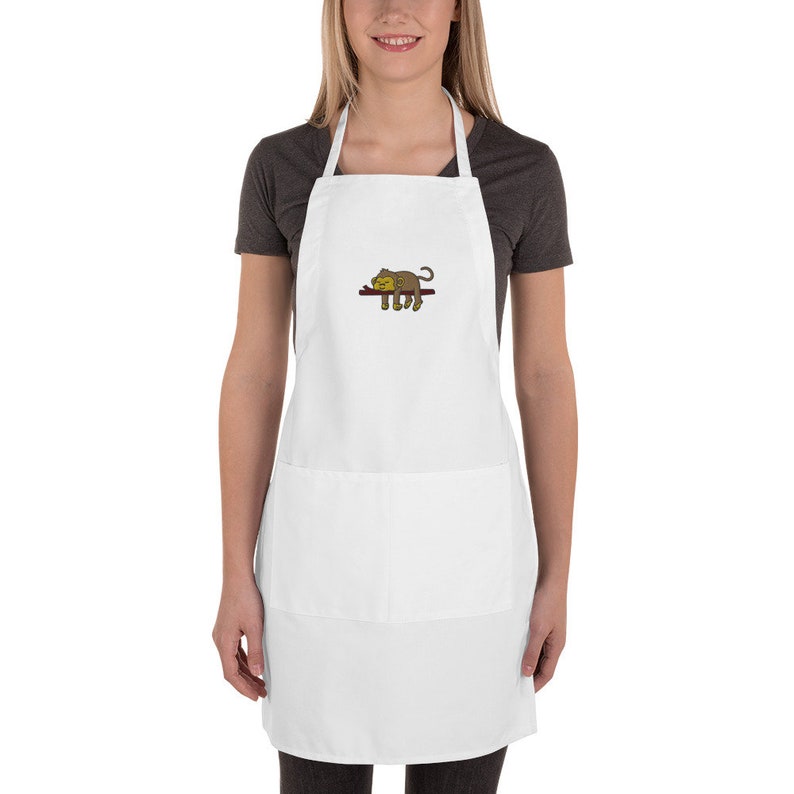 May include: White apron with a brown monkey sleeping on a branch embroidered on the front. The apron has a large pocket on the front.