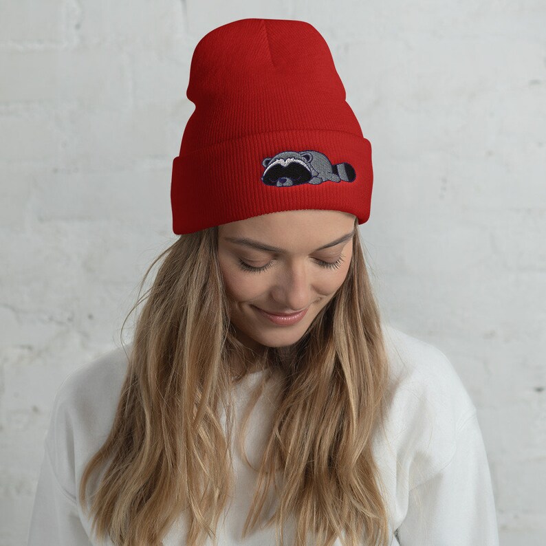 May include: A red beanie with a gray and black embroidered raccoon design. The raccoon is lying down with its head resting on its paws.