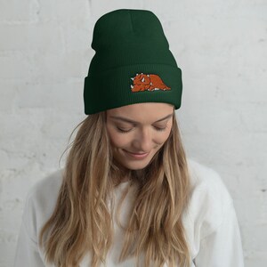 Puede incluir: Un gorro de lana verde oscuro con un dinosaurio marr&oacute;n bordado en la parte delantera.