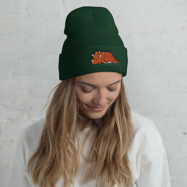 Puede incluir: Un gorro de lana verde oscuro con un dinosaurio marr&oacute;n bordado en la parte delantera.