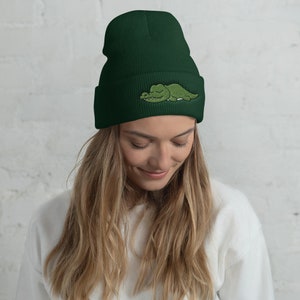 以下が含まれることがあります： 正面に緑色のワニが刺繍された濃い緑色のビーニー。ワニは目を閉じて横たわっています。