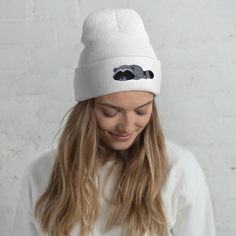 May include: White beanie with a black and gray embroidered raccoon design. The raccoon is lying down with its head resting on its paws.