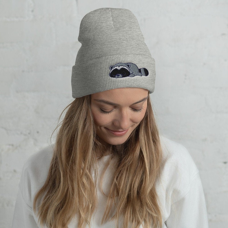 May include: A gray beanie with a black and white raccoon embroidered on the front. The raccoon is lying down with its head turned to the side.