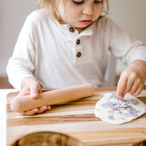 May include: A child is making a flower-shaped edible art project. They are using a rolling pin to flatten a white dough and pressing dried flowers into the dough. The dough is on a wooden cutting board. There are two coconut bowls with white dough in them.