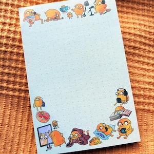 May include: A rectangular notepad with a light blue dotted grid pattern. The border features various illustrations of the orange dog character Jake from Adventure Time in different poses, including wearing headphones, reading, and using a computer.
