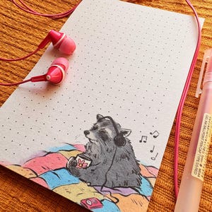 May include: A notebook with a dotted grid, featuring a raccoon wearing headphones and holding a cup, with musical notes. Pink earbuds and a pen are also present. The raccoon is sitting on a colorful patchwork blanket.