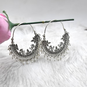 May include: A pair of silver-toned hoop earrings. Each earring features a semi-circular design with floral and bird motifs, and small dangling white beads. The earrings are displayed on a white, textured surface.