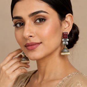 May include: A pair of drop earrings featuring a rectangular deep red stone set in oxidized silver, connected to a bell-shaped jhumka component with intricate metalwork and a fringe of small white beads at the base, worn on the earlobes.