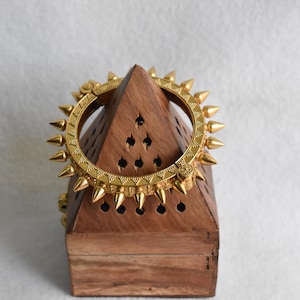 May include: A gold-coloured bangle bracelet with a spiked design rests on a wooden pyramid-shaped box. The bracelet features conical spikes around its circumference. The box has a natural wood grain finish.