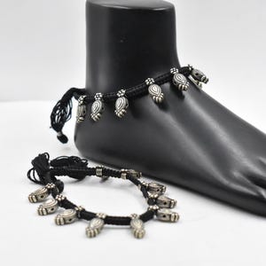 May include: Two black anklets with silver-colored charms. One anklet is on a black foot form, the other is lying on a white surface. The charms are teardrop-shaped with a detailed design. The anklets are tied with black cords.