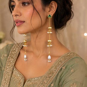 May include: Long gold-tone drop earrings featuring a green leaf-shaped stud, two spherical gold beads with small white pearl accents, and a single white teardrop-shaped pearl at the base, suspended from a thin gold chain.