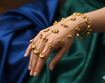 May include: A gold-toned hand chain bracelet featuring a series of delicate chains connected to a wrist cuff and finger rings. The design incorporates small crescent moon charms and clusters of tiny spherical bells suspended along the thin metal chains.
