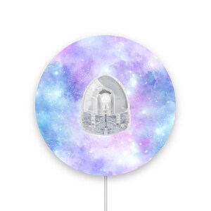 Op de afbeelding: Een ronde, witte, wandgemonteerde nachtlamp met een galaxy-thema. De nachtlamp heeft een blauw, paars en roze galaxy-patroon met witte sterren.
