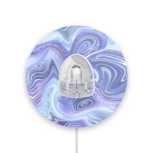 Peut inclure: Une plaque murale ronde et blanche avec un motif marbré bleu et violet. La plaque a un cache-prise rond et blanc en plastique au centre.