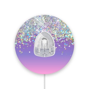 Può includere: Un gancio adesivo da parete rotondo con uno sfondo sfumato rosa e viola. Il gancio è ricoperto di glitter iridescente e ha un gancio in plastica trasparente.