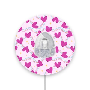 May include: A round, pink and white sticker with a heart pattern and rainbow accents. The sticker has a clear, round opening in the center.