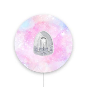 May include: A clear, egg-shaped device with a grey top, set against a circular background of pink, purple, and blue watercolour-style patterns. A thin white cord extends from the bottom.