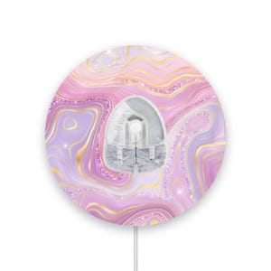 Peut inclure: Une poignée de téléphone ronde de style agate rose et violette avec des accents dorés et des paillettes. La poignée est attachée à un cordon blanc.