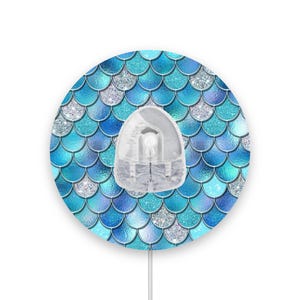 Op de afbeelding: Een ronde wanddecoratie met een blauw en zilveren zeemeerminschubbenpatroon. De decoratie heeft een witte, ronde, plastic stop in het midden.